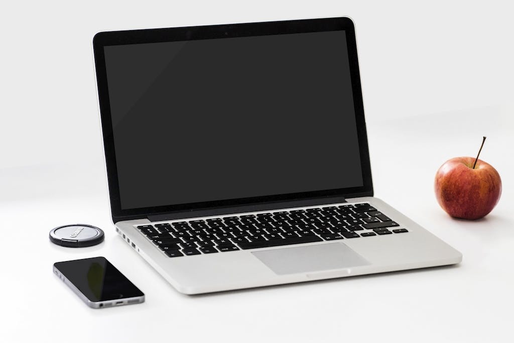 A minimalist workspace featuring a laptop, smartphone, and an apple on a clean white desk.