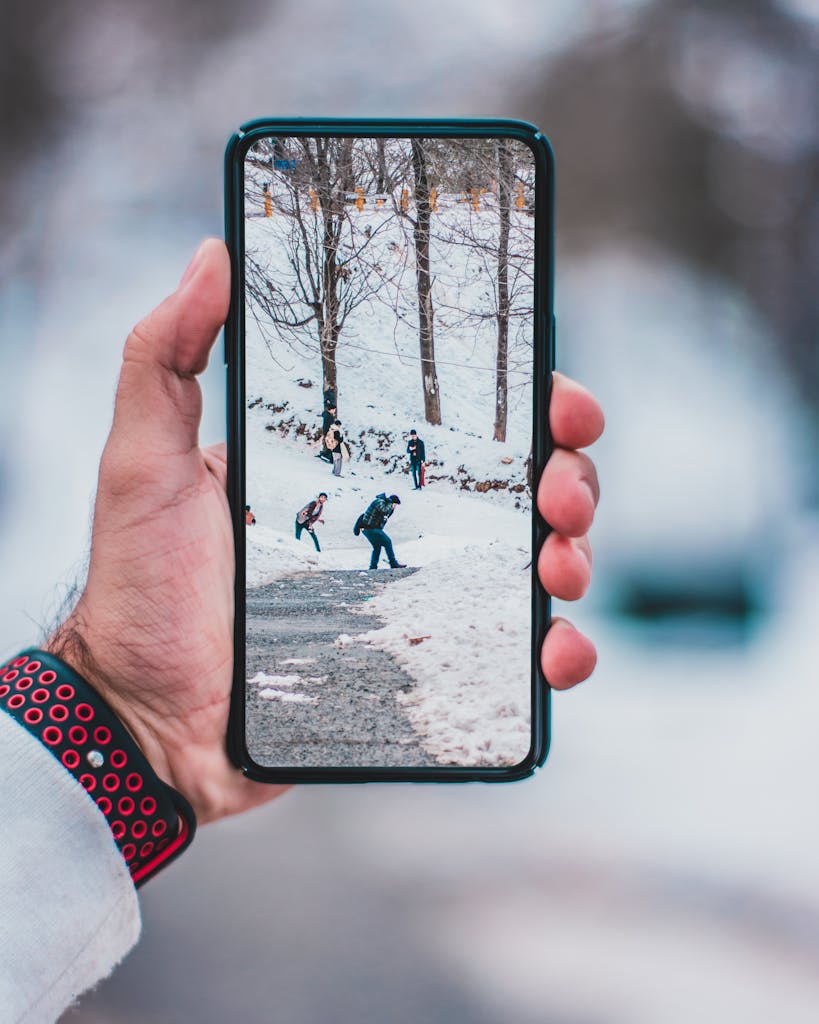 Hand holding smartphone displaying a winter scene with people walking on snow-covered path.